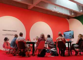A photo of 9 participants taking part in a Feminist School of Painting workshop. There is a bright red carpet on the floor. Participants sit around four tables, each with space for four chairs. To the left, is a screen. The wall is bright orange.