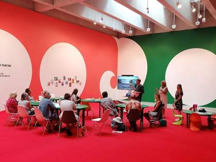 A photo of 9 participants taking part in a Feminist School of Painting workshop. There is a bright red carpet. Participants sit around five tables. To the left of centre, is a screen showing a landscape.