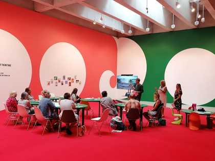 A photo of 9 participants taking part in a Feminist School of Painting workshop. There is a bright red carpet. Participants sit around five tables. To the left of centre, is a screen showing a landscape.