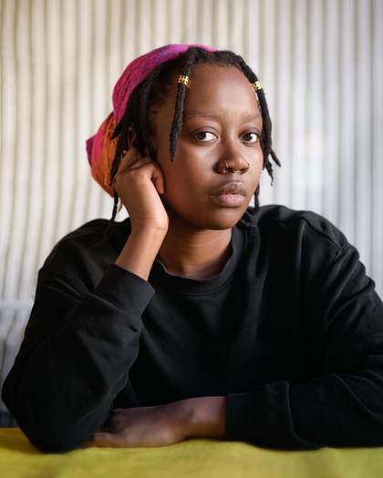 A person wearing a pink headscarf and a black sweater, resting their head on their hand while looking directly at the camera indoors.