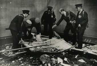 Black and white photo of a group of men inspecting bomb damage on the floor of a gallery, there is a painting on the floor.