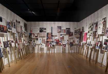A room with white walls and wooden floor. The three walls of the room visible have placards of different sizes and heights visible. The placards have images of women from magazines on them.