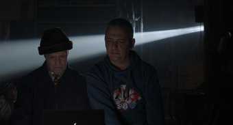 Two men sit in a darkened room with light streaming in possibly from a projector
