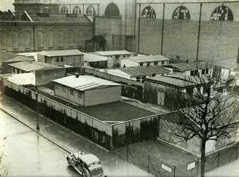 Black and white photo of temporary buildings in Tate Britain grounds.  There is a car in the foreground.