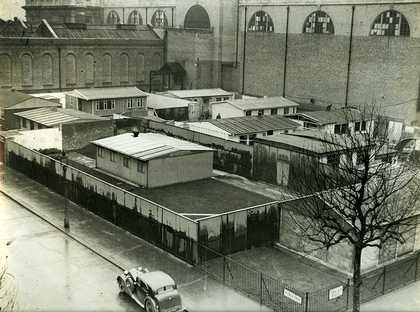 Black and white photo of temporary buildings in Tate Britain grounds. There is a car in the foreground.