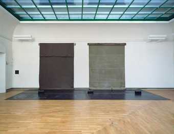 Photograph of a large installation by Joseph Beuys in the Staatsgalerie Stuttgart, Germany. Two tarpaulin-like objects – one made of rubber, the other felt – are installed on the wall and touch down to the floor in a thick, heavy bulge.