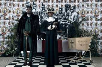 A Black man and woman in expensive looking, early twentieth century British style clothing. Behind them is an altar, above which is a photo of a group of Black men wearing indigenous clothing. A shopping trolley bears the words ‘Shopping For Jesus’.