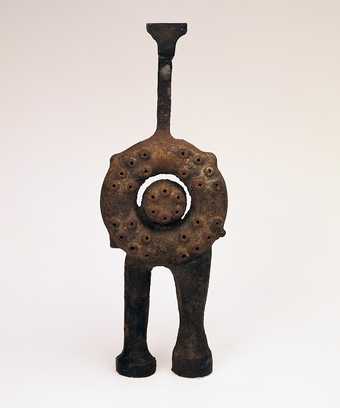 Abstract sculpture made of brown rusty metal, standing on two animal-like legs, above which is a circle form contained within another circle and above a long funnel-shaped form. The circular forms have a series of circular holes in them.