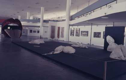 Large exhibition space on two levels with white walls and columns, pictures on the walls, objects wrapped in cloth, a large sculpture of a mouth with protruding tongue