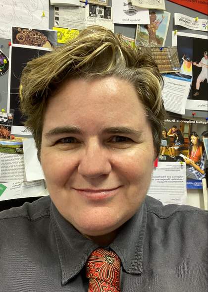 Portrait of Sandy O’Sullivan sitting in an office in front of a wall with lots of pictures, wearing a grey shirt and red pattern tie.