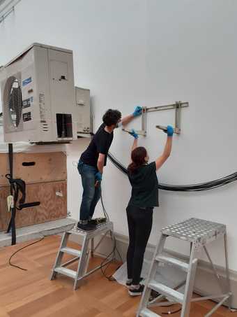 Two people hold a bracket up against a gallery wall, one is up a step ladder