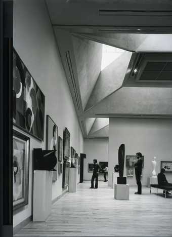 Black and white image of a gallery with sculptures and paintings in a modern looking gallery.  There are a couple of people looking at the artworks.