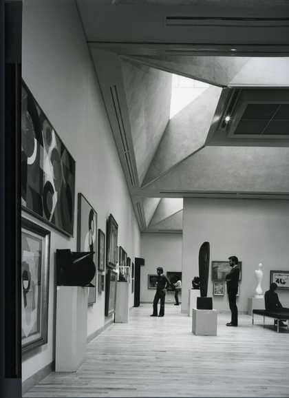 Black and white image of a gallery with sculptures and paintings in a modern looking gallery. There are a couple of people looking at the artworks.