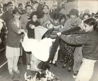 A black-and-white photo of a group of people, a small fire in the foreground over which women hold pieces of cloth