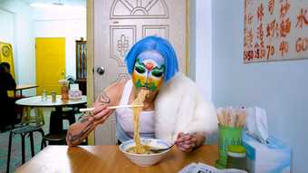 A person sits eating noodles in a cafe, chopsticks and spoon in either hand. They wear a white vest with a white fur draped over one shoulder, bright blue short hair and their face is painted with an abstract landscape, a lotus flower between the eyes.