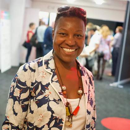 Portrait of Veronica McKenzie at an event, wearing a floral jacket, large beaded necklace and sunglasses on her head.
