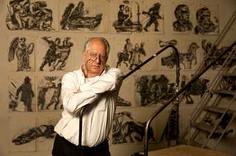 Portrait of William Kentridge standing in front of some figurative drawings on the wall