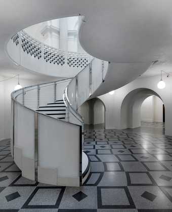 Photo of a circular staircase rising out of a patterned stone floor with a white ceiling above.