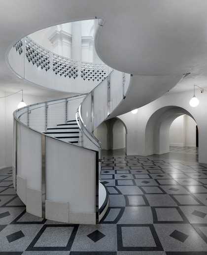 Photo of a circular staircase rising out of a patterned stone floor with a white ceiling above.