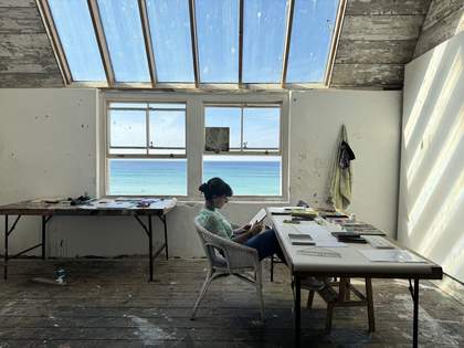 The artist sits reading at a table in her studio in St Ives. There is a window behind looking out to sea and the studio is lit with natural light from the large skylights above her.