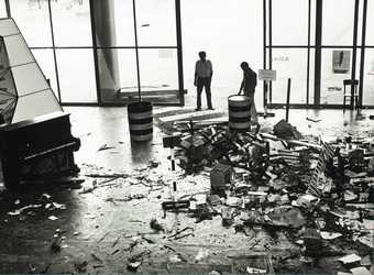 Two people stand in front of large glass doors, looking at the debris strewn across the pedestrian crossing, including signage, bottles, food, packaging, printed matter and blood