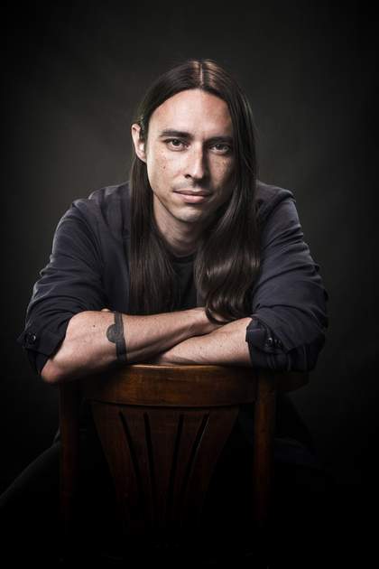 Portrait of Joseph M. Pierce sitting backwards on a chair in front of a black background, wearing a black shirt. 