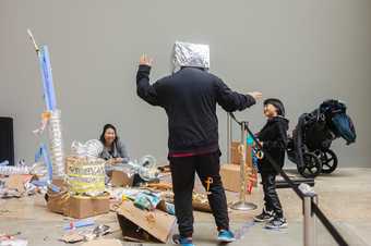 A man has a tin foil covered cardboard box on his head and two people look on smiling