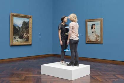 Installation view of two women standing on a plinth with bottles held between them