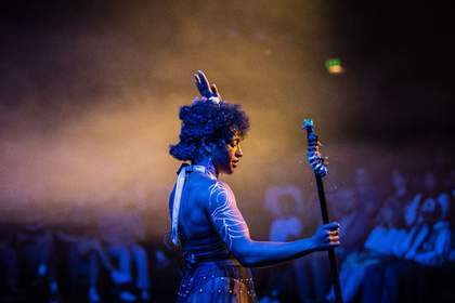 A photograph of a performer on stage bathed in blue and yellow light.