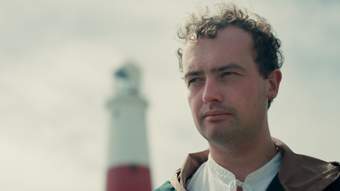 Person with curly short hair looking off into the background with an out of focus lighthouse behind them