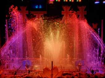 film still from the watershow extravaganza. shoots of water brightly lit with pink and red lighting