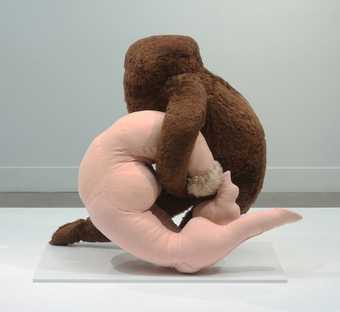 This is a photograph of one of Dorothea Tanning's soft sculptures on display in a white-walled gallery space. One sculpture is light pink and fleshy and is intertwined with a brown, hairy figure.