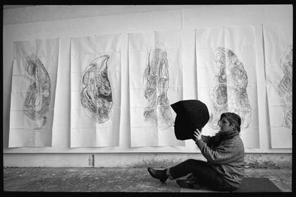 The artist is sat on the floor of her studio in St Ives, working with paper cut outs. Behind her large drawings on paper are hung on the wall.