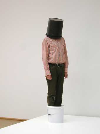 Installation shot of a person standing on a plinth in a bucket with a bucket on his head