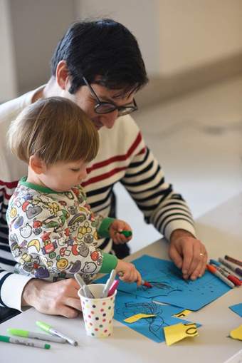A photograph of an adult sat with a child who is drawing on some paper