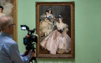 A person with a professional camera filming an artwork by John Singer Sargent against a bright green wall