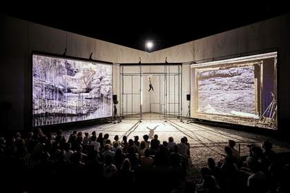A crowd of people watching a performance involving a person hanging by their arms from a large metal structure in between two large screens