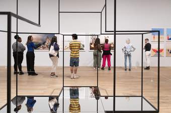 Behind a metal frame structure, a group of people look at art on the wall.