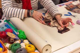 A teacher sticks down a printed photograph of a woman onto a large roll of paper, holding scissors and colourful sellotape in her hands as she does. Around the paper are many colourful balls of string and other art materials, ready to be used in the ac