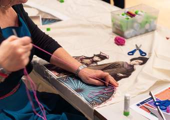 A teacher sews a printed photograph of a colourful pattern onto a sheet of fabric. Other art materials are also on the table, including a box full of glue sticks and some pairs of safety scissors.