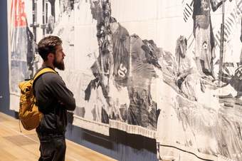 A teacher with a yellow rucksack looks at Zohra Opoku’s ‘Queens and Kings’ in the gallery, a large-scale tapestry with greyscale photographs printed onto it.