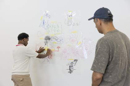 In the right-hand corner of the image, artist Abbas Zahedi stands with their back to the camera, observing a young person as they arrange drawings made on acetate on a wall.