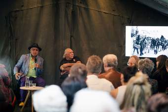 An audience facing two male speakers, one with a microphone and there is a screen to the side.