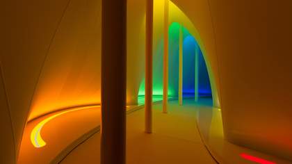 An interior walkway with curved walls and columns that is illuminated by gradient lighting, transitioning from warm orange and yellow in the foreground to green and deep blue in the distance.