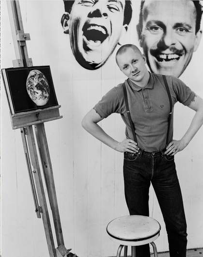 A black and white photograph of Andrew Heard. He has a shaved head and wears a Fred Perry polo shirt. He poses with his hands on his hips next to a book placed on an easel. The book shows a photo of the globe.