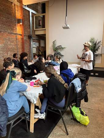 A photograph of a person talking to a group of young people who are sat round a table.