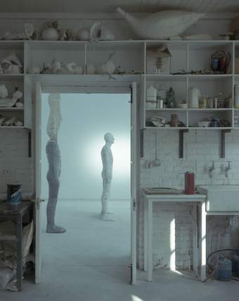 A photograph inside an artist studio looking through a doorway towards two human-like sculptures.