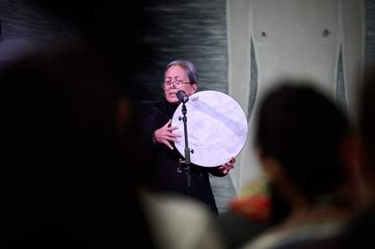 A woman stands in front of a microphone in a dark room, lit by a spotlight, her eyes closed, holding a circular drum in her hand