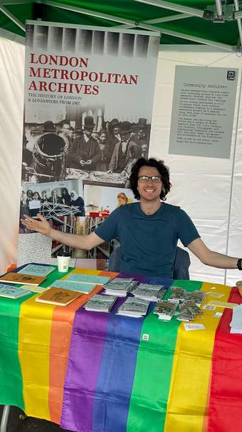A smiling person with long hair and glasses sits at a table. The table has a rainbow-coloured covering and has papers and leaflets on it.  In the background there is a banner that says London Metropolitan Archives and poster that says community archives