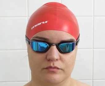 A headshot of a person in a red swimming cap and goggle, standing against a white tiled wall.